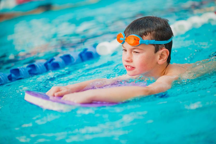 Junge mit orangenen Schwimmbrille übt auf einem Schwimmbrett im Wasser eines Schwimmbeckens.
