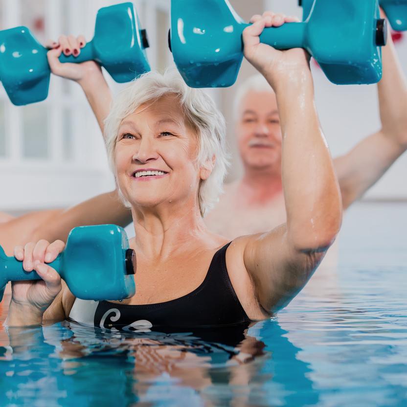 Drei Personen im Wasser heben blaue Hanteln, lächeln und üben Aquafitness in einem hellen Schwimmbad.